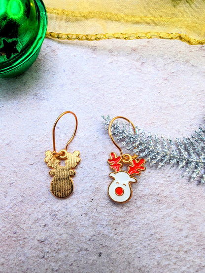 The Rosie Reindeer Hoop Earrings by Silver Sunbird showcase a reindeer-themed design on a textured surface. One earring highlights a simple gold silhouette, while the other displays an enamel white face with red antlers. A green ornament and silver pine branch in the background enhance the festive charm of these rose gold treasures.