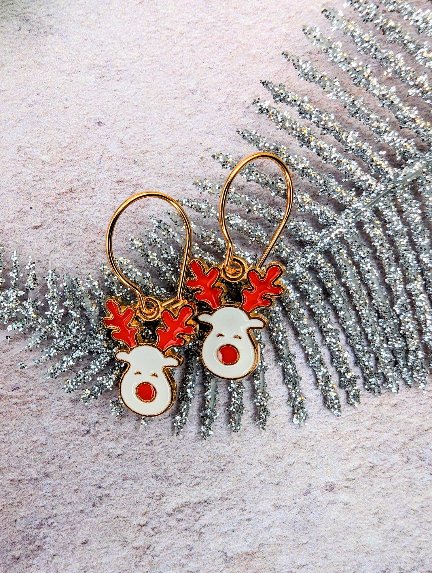 Rosie Reindeer Hoop Earrings by Silver Sunbird, adorned with festive enamel designs of red antlers and noses, are beautifully showcased on a glittery silver fern-like decoration against a lightly textured backdrop.
