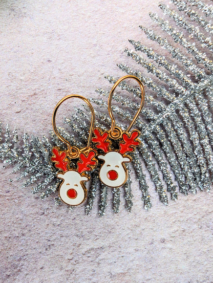 Rosie Reindeer Hoop Earrings by Silver Sunbird, adorned with festive enamel designs of red antlers and noses, are beautifully showcased on a glittery silver fern-like decoration against a lightly textured backdrop.