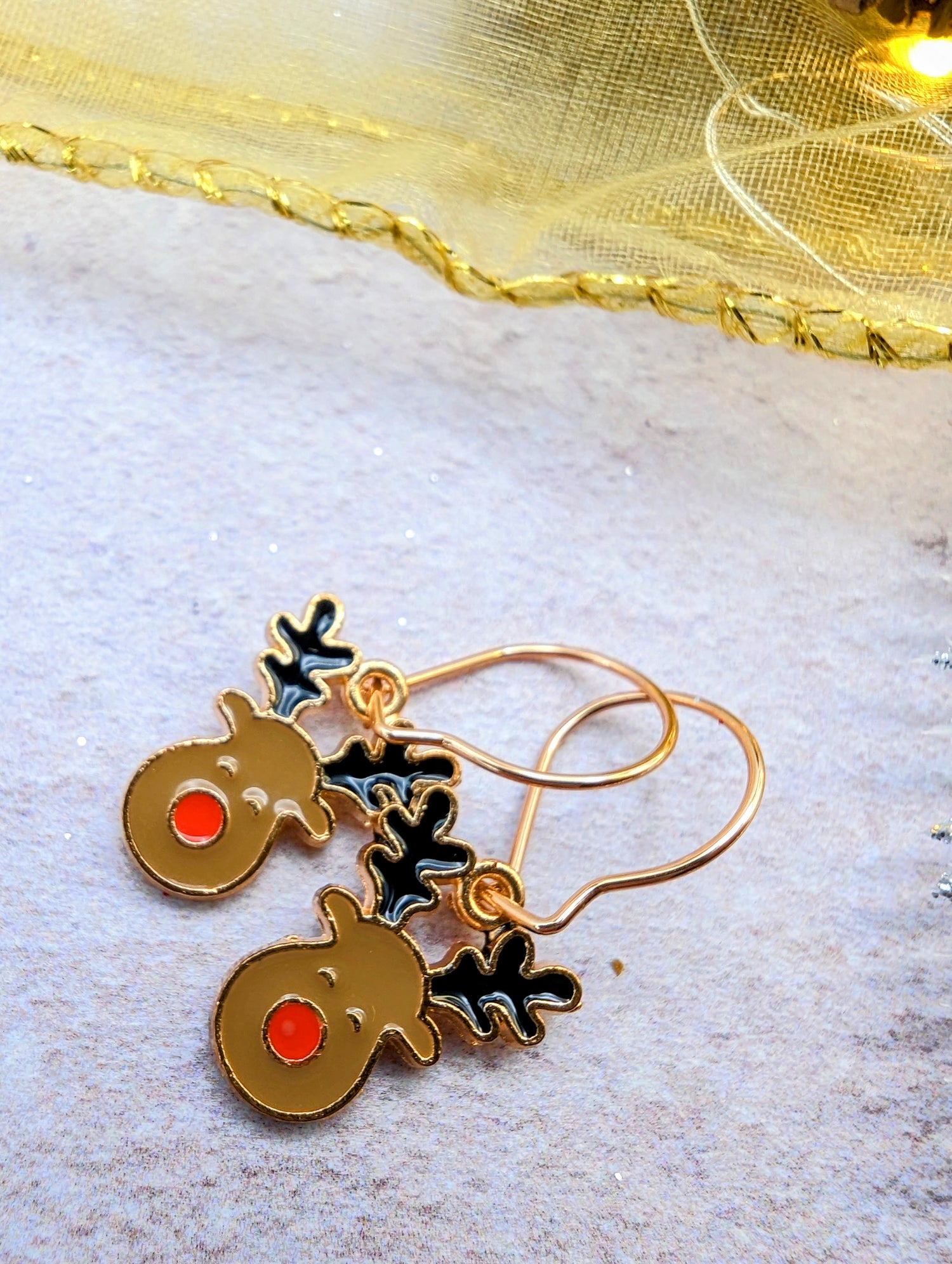 A pair of Rosie Reindeer Hoop Earrings by Silver Sunbird rests on a textured silver surface. The reindeer faces are brown adorned with red noses and black antlers, enhanced by a sheer, shimmery golden fabric in the background.