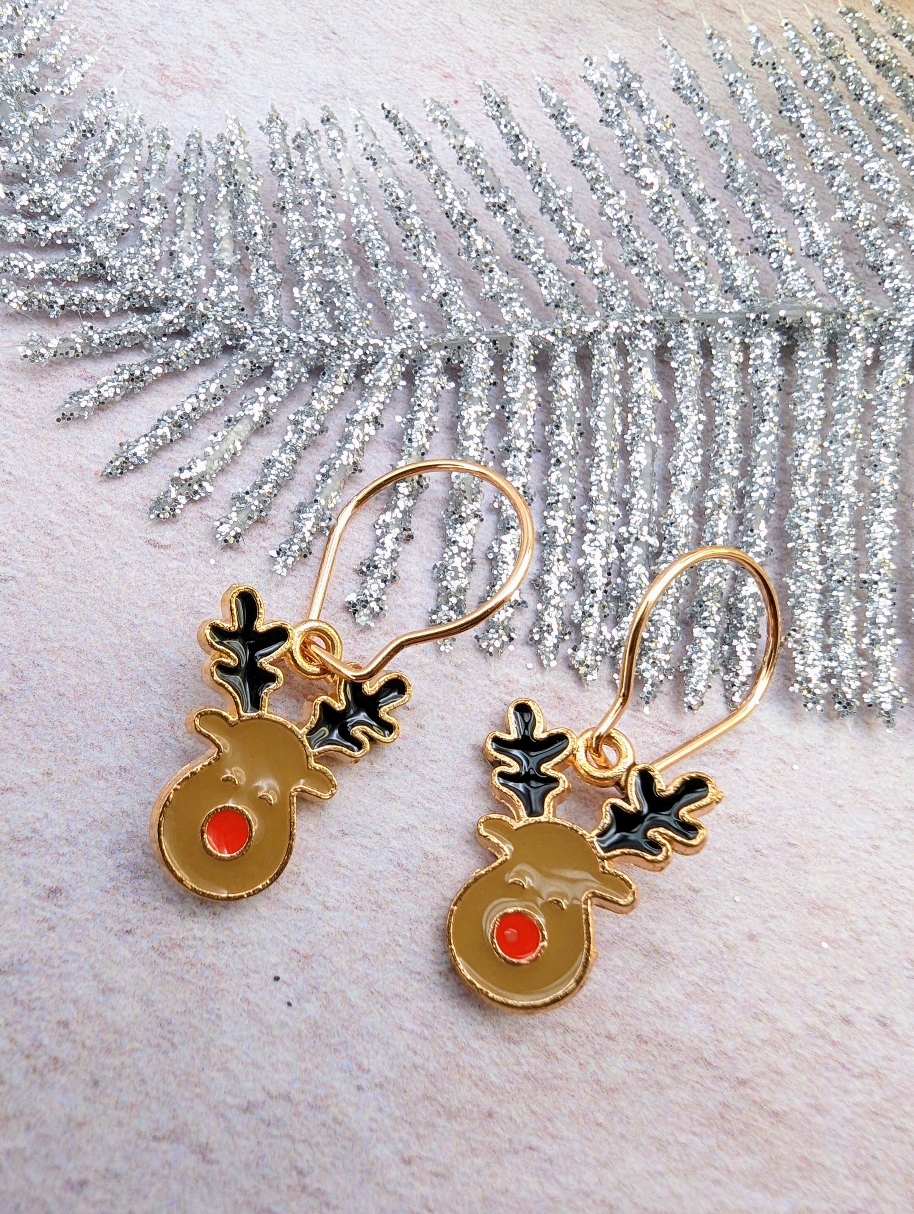 The Rosie Reindeer Hoop Earrings by Silver Sunbird, featuring gold bodies, black antlers, and red noses, dangle elegantly from a sparkling silver branch. This festive enamel design perfectly captures the holiday spirit.
