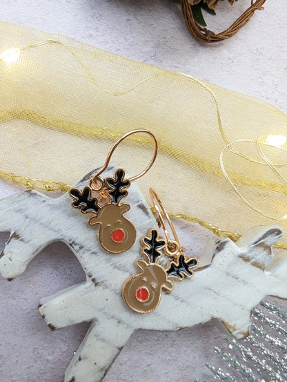 The Rosie Reindeer Hoop Earrings by Silver Sunbird are showcased on a rustic white wooden background. Featuring black antlers and cheerful red noses, these festive enamel earrings are accented with a sheer yellow ribbon and small lights, exuding a joyful holiday charm.