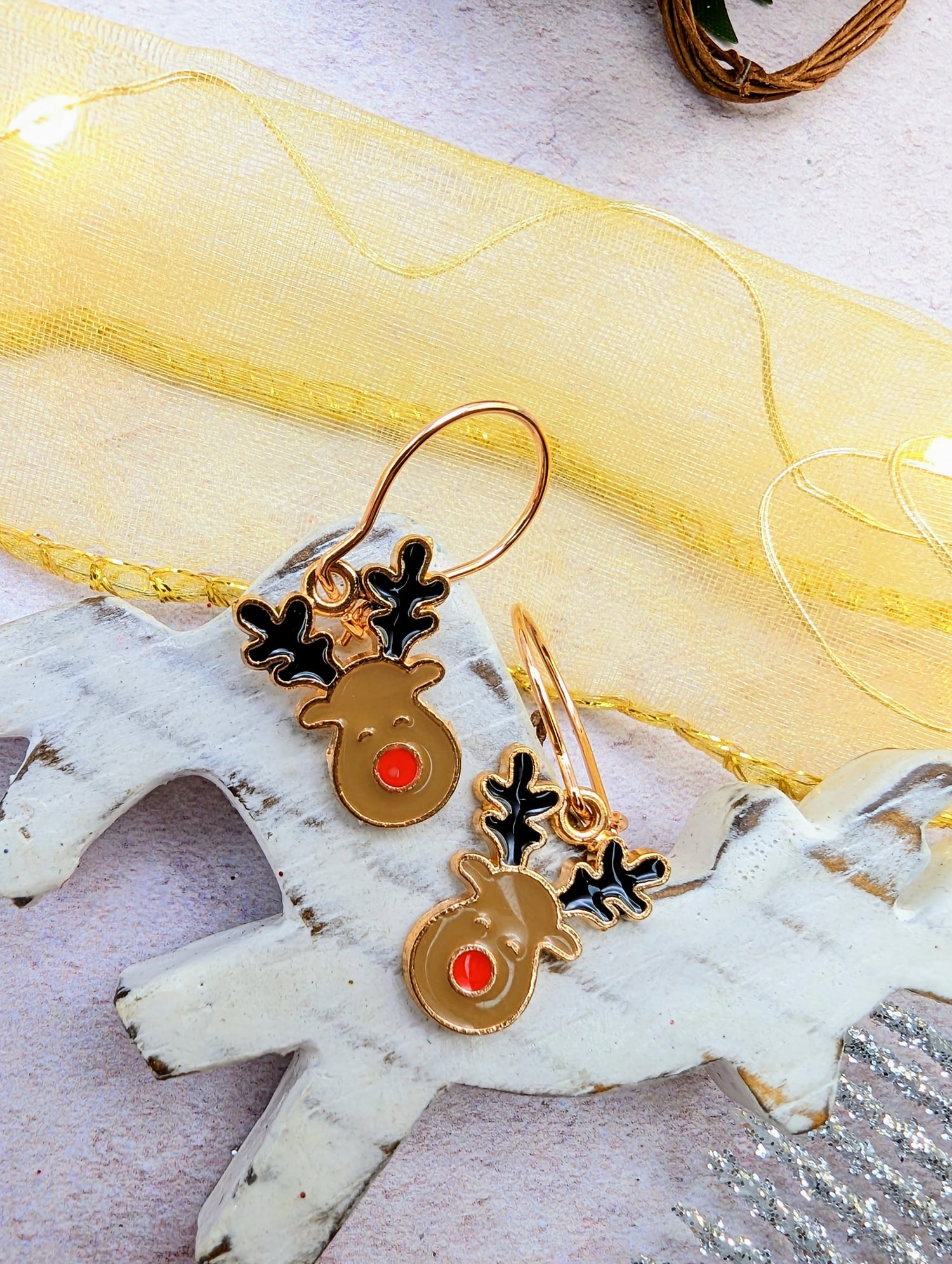 The Rosie Reindeer Hoop Earrings by Silver Sunbird, featuring black antlers and red noses, are elegantly displayed on a white wooden reindeer figure. A sheer yellow ribbon and small string lights create a festive backdrop, making these charming enamel earrings the ideal seasonal accessory.