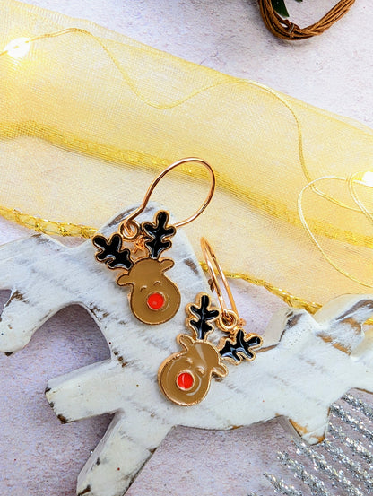 The Rosie Reindeer Hoop Earrings by Silver Sunbird, featuring black antlers and red noses, are elegantly displayed on a white wooden reindeer figure. A sheer yellow ribbon and small string lights create a festive backdrop, making these charming enamel earrings the ideal seasonal accessory.