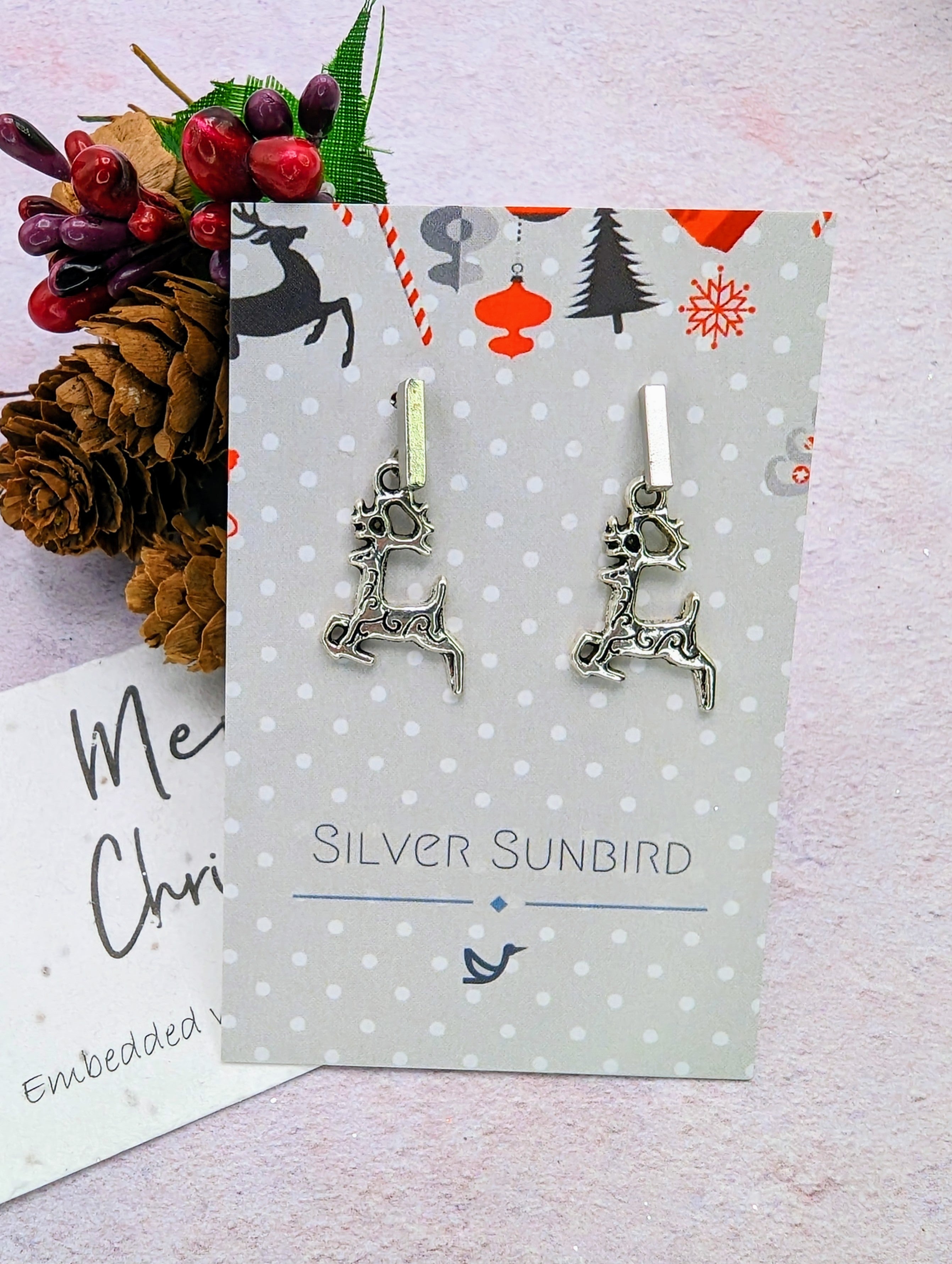 The Prancing Reindeer Earrings by Silver Sunbird are presented on a decorative card embellished with polka dots and Christmas-themed illustrations. Pine cones and festive berries provide a cheerful backdrop, accentuating the handmade jewelry&