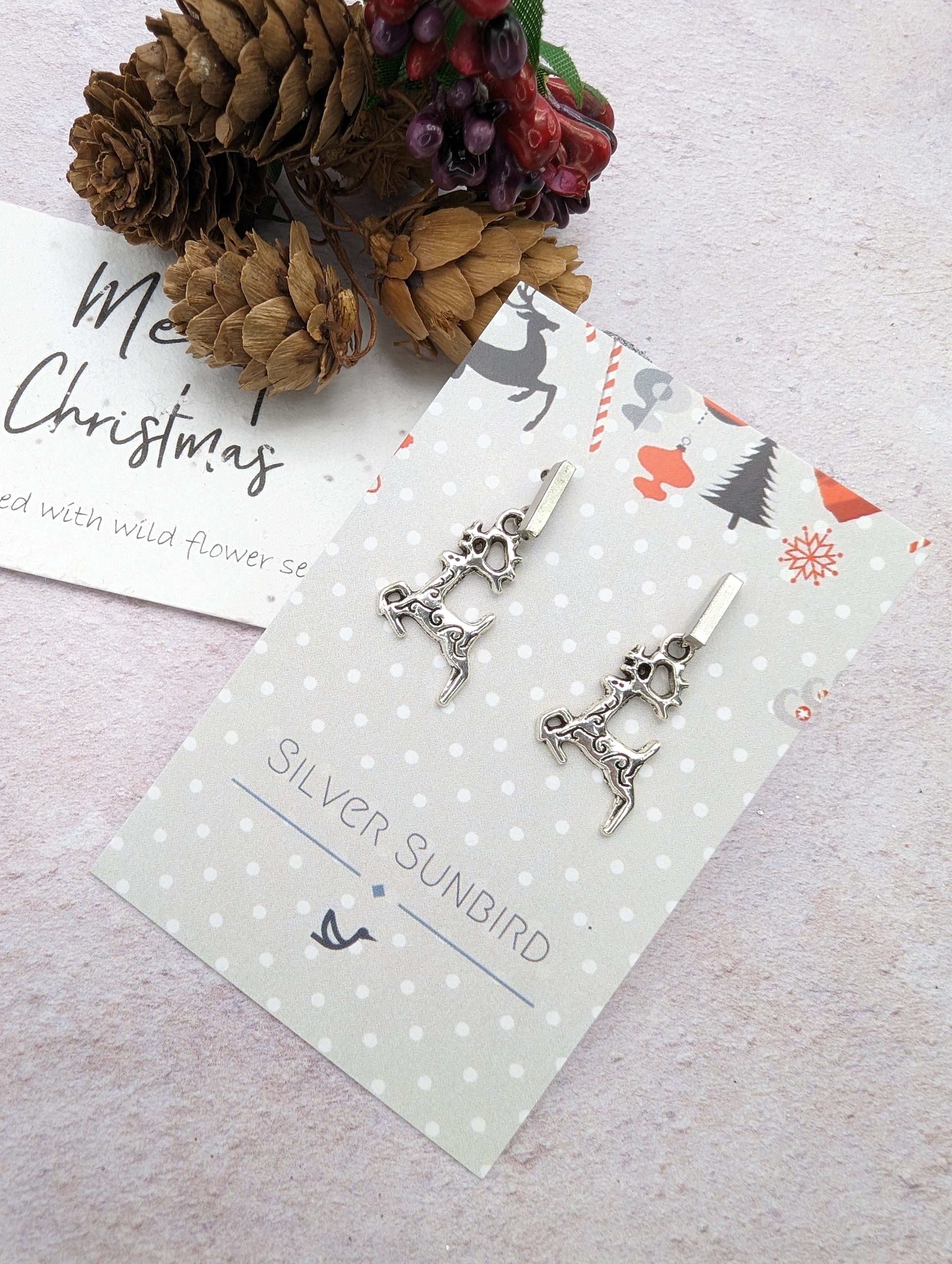The &quot;Prancing Reindeer Earrings&quot; by Silver Sunbird are crafted from stainless steel and elegantly displayed on a decorative card featuring a festive design. The set includes a &quot;Merry Christmas&quot; note and is adorned with dried pinecones and berries against a light background, creating delightful handmade jewelry perfect for the season.