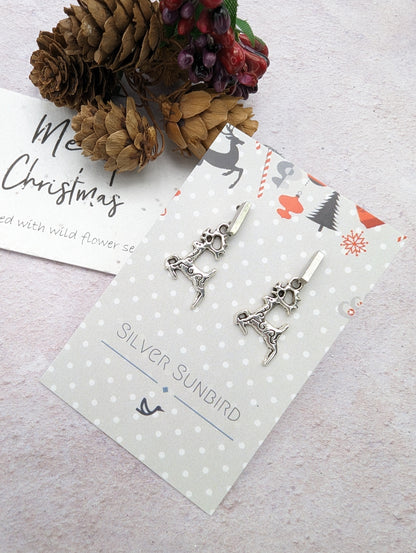 The &quot;Prancing Reindeer Earrings&quot; by Silver Sunbird are crafted from stainless steel and elegantly displayed on a decorative card featuring a festive design. The set includes a &quot;Merry Christmas&quot; note and is adorned with dried pinecones and berries against a light background, creating delightful handmade jewelry perfect for the season.