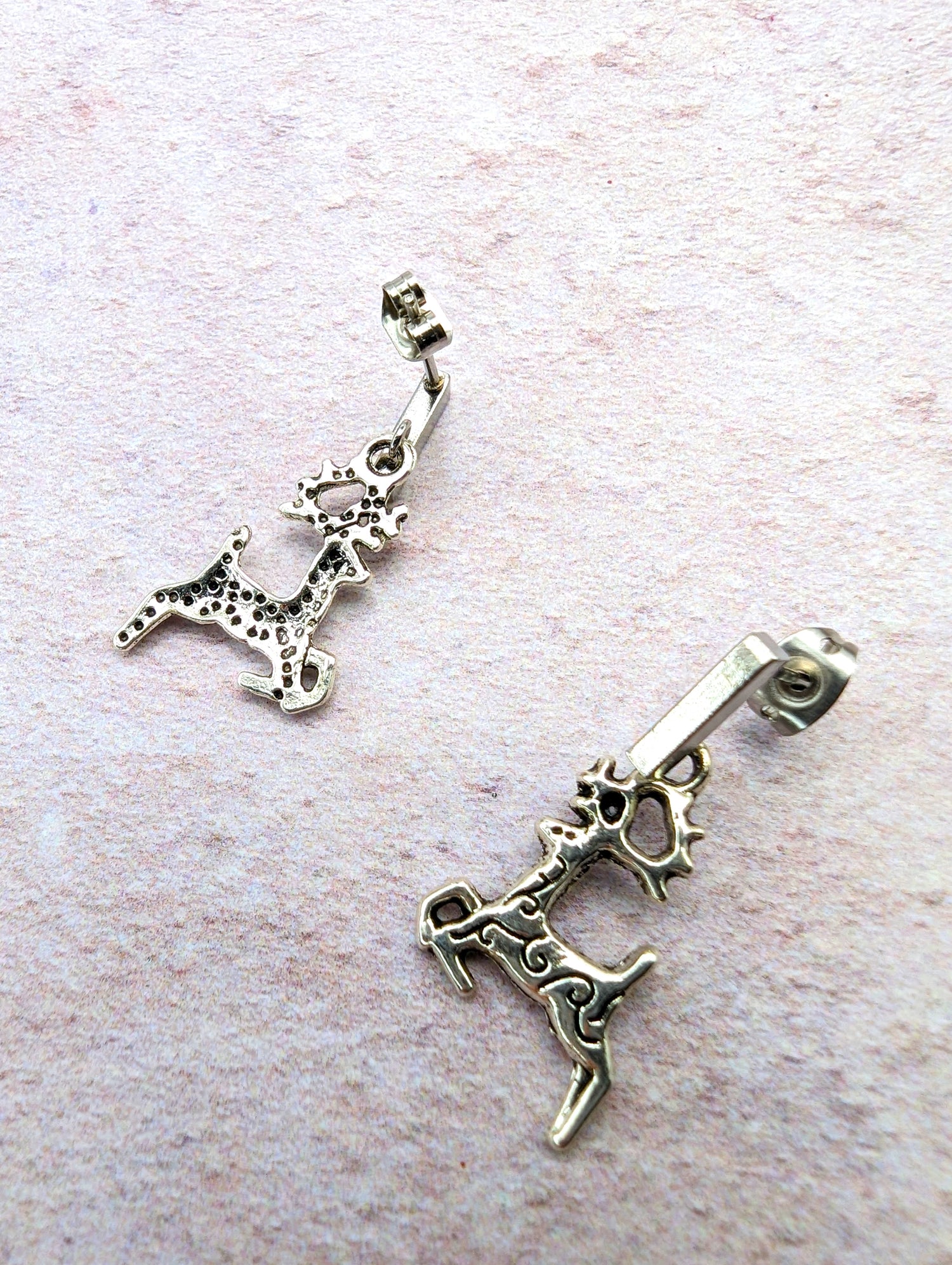 A pair of Prancing Reindeer Earrings from Silver Sunbird, crafted from stainless steel with a rustic, antique finish and intricate detailing, lies on a textured, light-colored surface. These playful and dynamic earrings add charm to your collection of handmade jewelry.