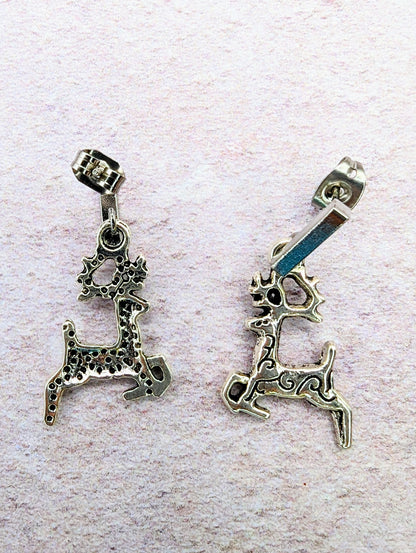 On a textured light pink surface, two Prancing Reindeer Earrings from Silver Sunbird are displayed. Each handmade silver earring showcases unique and intricate patterns, with one featuring a dotted design and the other a swirled pattern. Both pieces include antlers and a bail on top for hanging, highlighting exquisite craftsmanship in jewelry design.