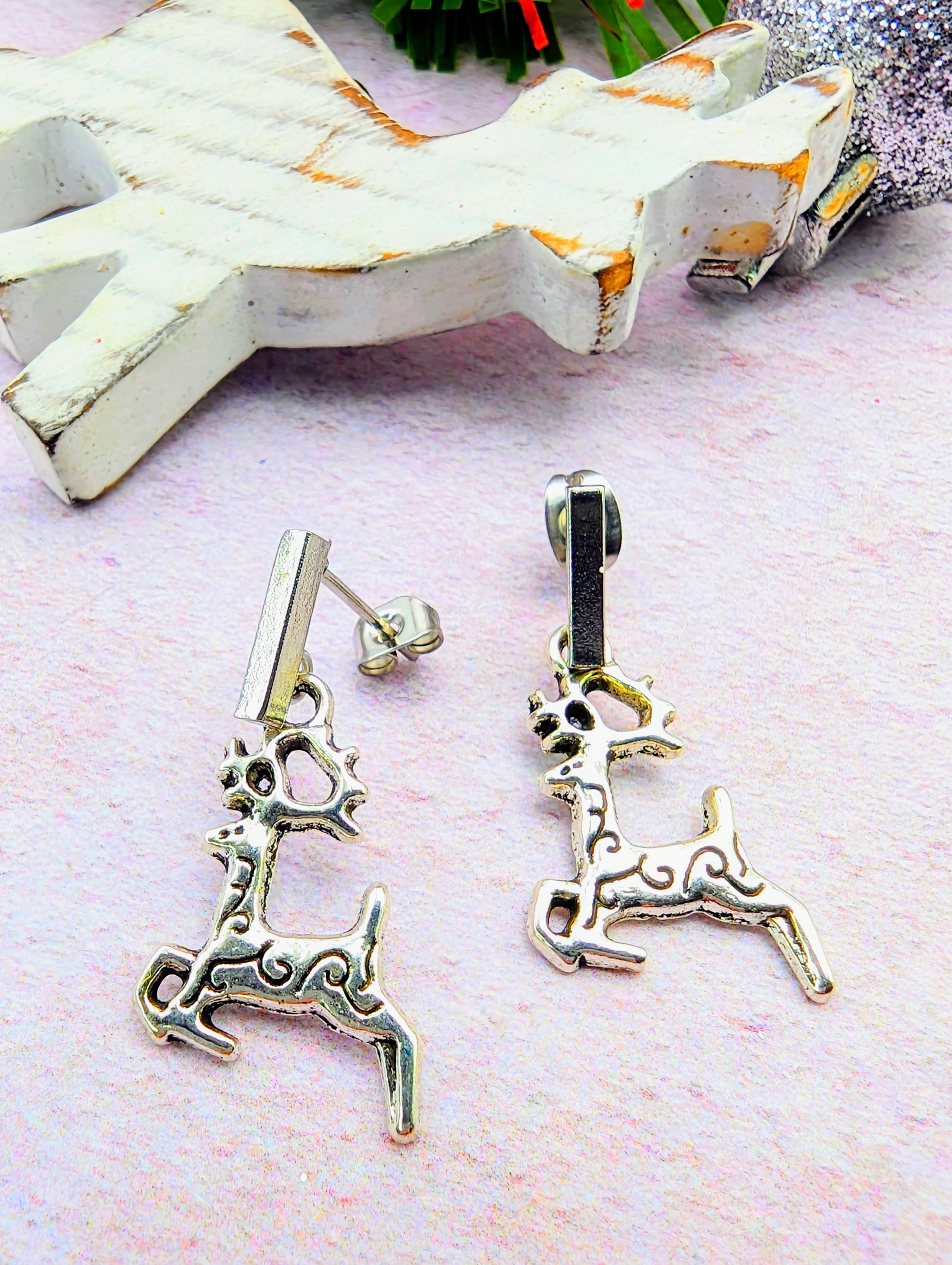 A pair of Prancing Reindeer Earrings by Silver Sunbird rests on a light surface, complemented by a wooden reindeer decor in the background. These exquisite handmade jewelry pieces boast intricate, artistic designs and are elegantly styled, making them the perfect reindeer charms for any occasion.