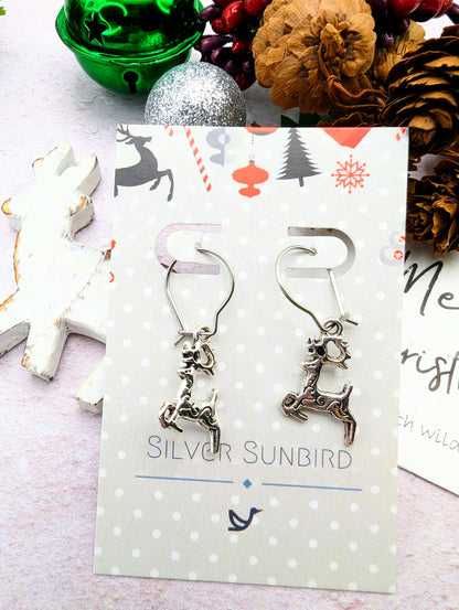 The Prancing Reindeer Earrings by Silver Sunbird are presented on a celebratory card featuring a Christmas motif. The backdrop showcases a green bauble, pinecone, and red berries for a festive theme. Crafted from stainless steel, these earrings add an element of handmade jewelry elegance to any holiday ensemble.
