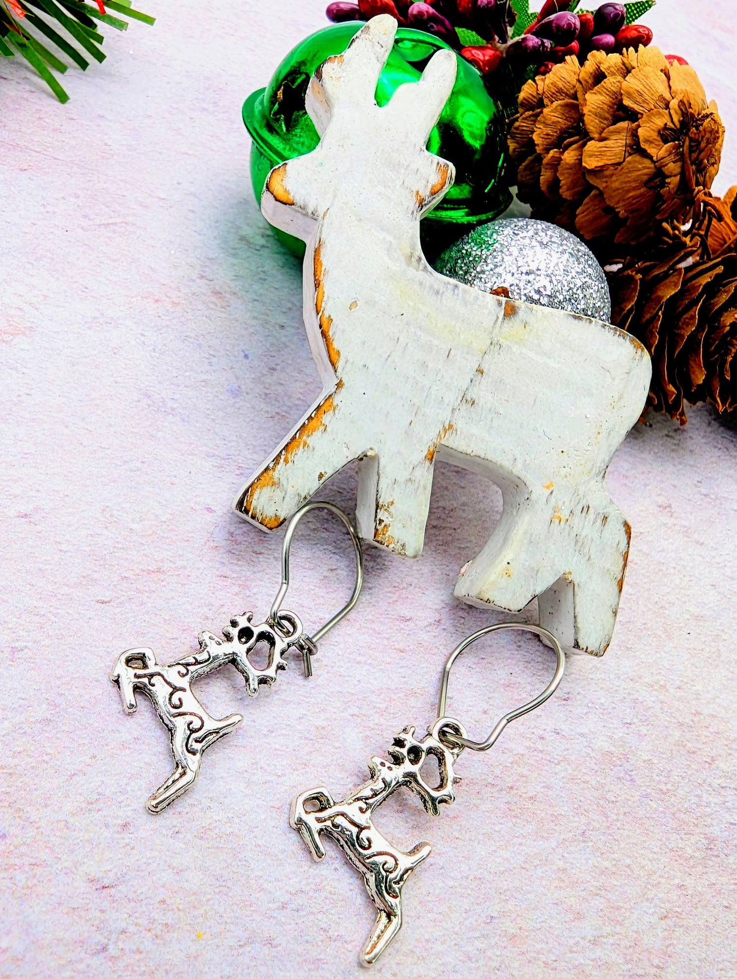 Festive display showcasing a wooden reindeer figure adorned with pine cones, and green and silver ornaments. Below, the Prancing Reindeer Earrings by Silver Sunbird are elegantly placed on a soft pink surface.