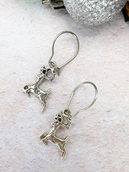 Prancing Reindeer Earrings by Silver Sunbird are displayed on a textured surface alongside a sparkling silver ornament.
