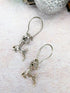 Prancing Reindeer Earrings by Silver Sunbird are displayed on a textured surface alongside a sparkling silver ornament.