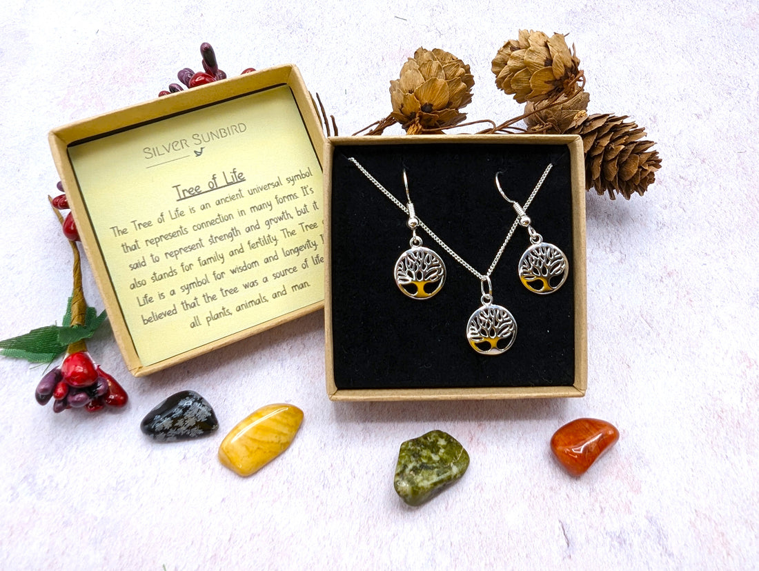 The Silver Sunbird Sterling Silver Tree of Life Jewellery Set includes a necklace and hypoallergenic earrings in a black box with an explanation card, pine cones, red berries, green leaves, and five colorful stones.