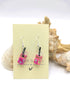 The Party Cocktail Earrings by Silver Sunbird, featuring pink and clear resin with white flowers and black strips, dangle from a "Sugar Sugar" card. Perfect for summer, they sit beautifully against seashell-adorned white surfaces.