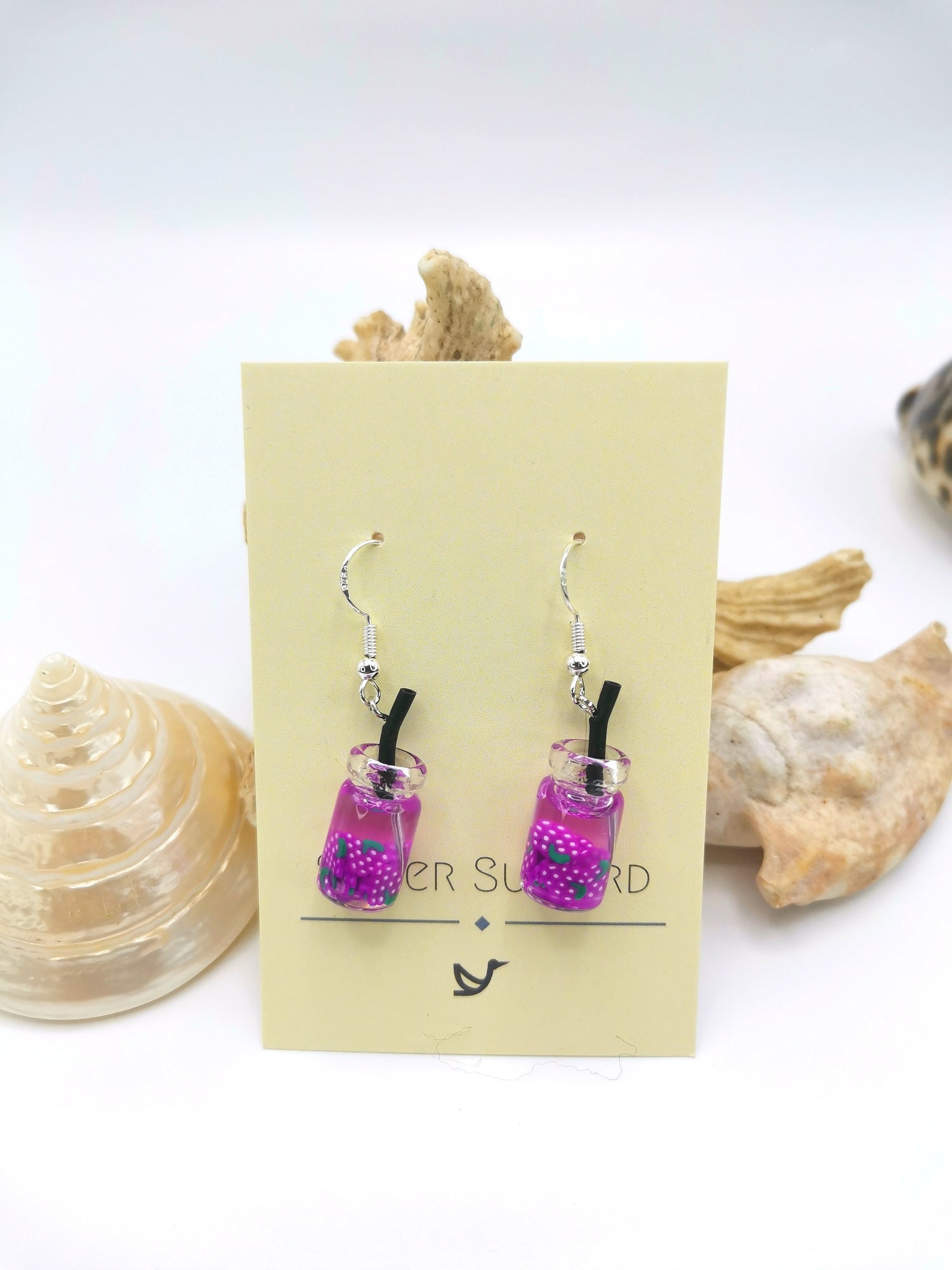 The Silver Sunbird Party Cocktail Earrings are handmade to look like tiny purple beverage bottles with straws and colorful beads. Displayed on a card amidst seashells, they embody the playful spirit of summer resin fruits.