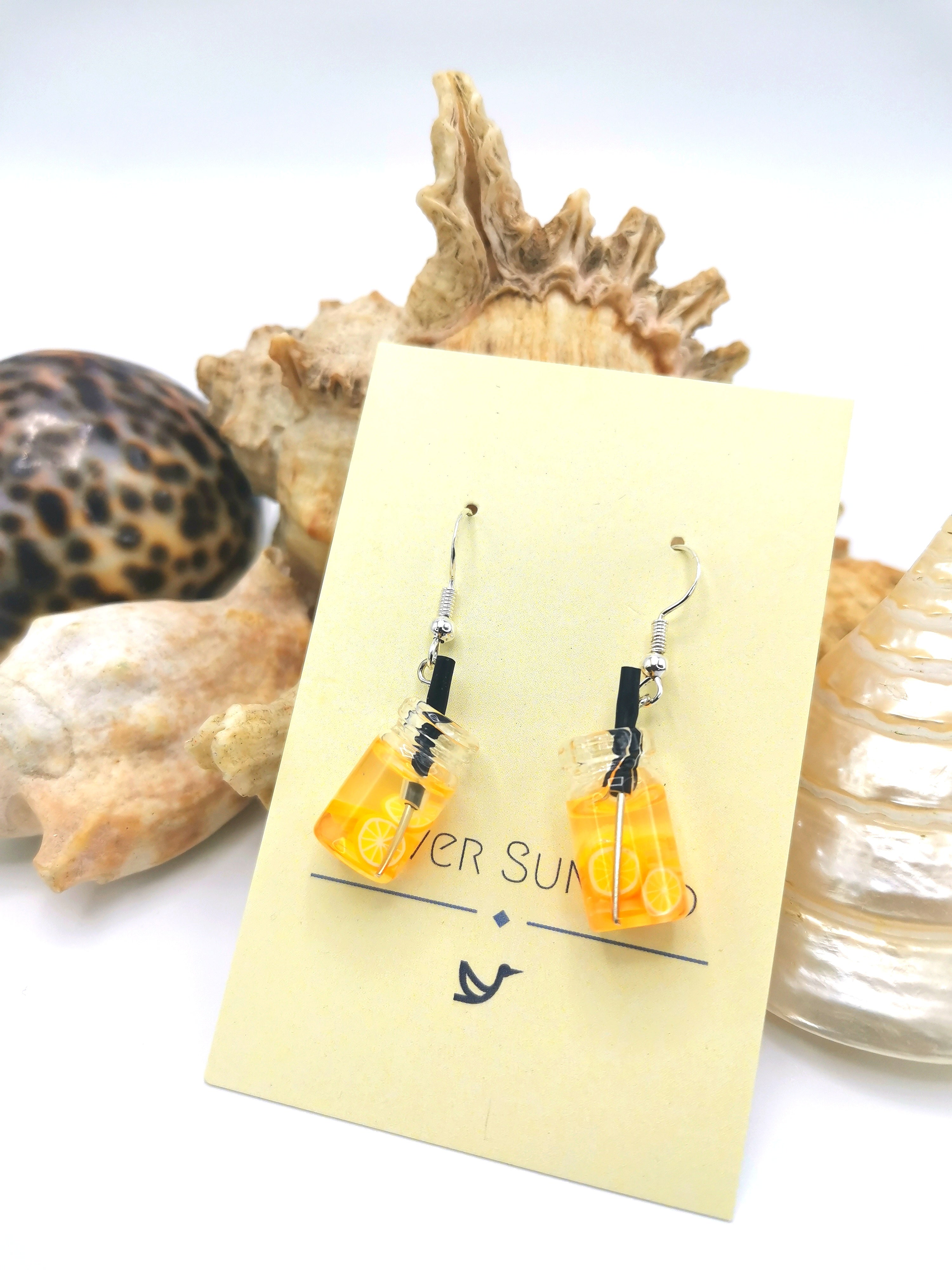 The Party Cocktail Earrings by Silver Sunbird are handmade with lever-back hooks, featuring miniature beaker designs filled with yellow liquid. Displayed against seashells, they evoke the essence of summer.