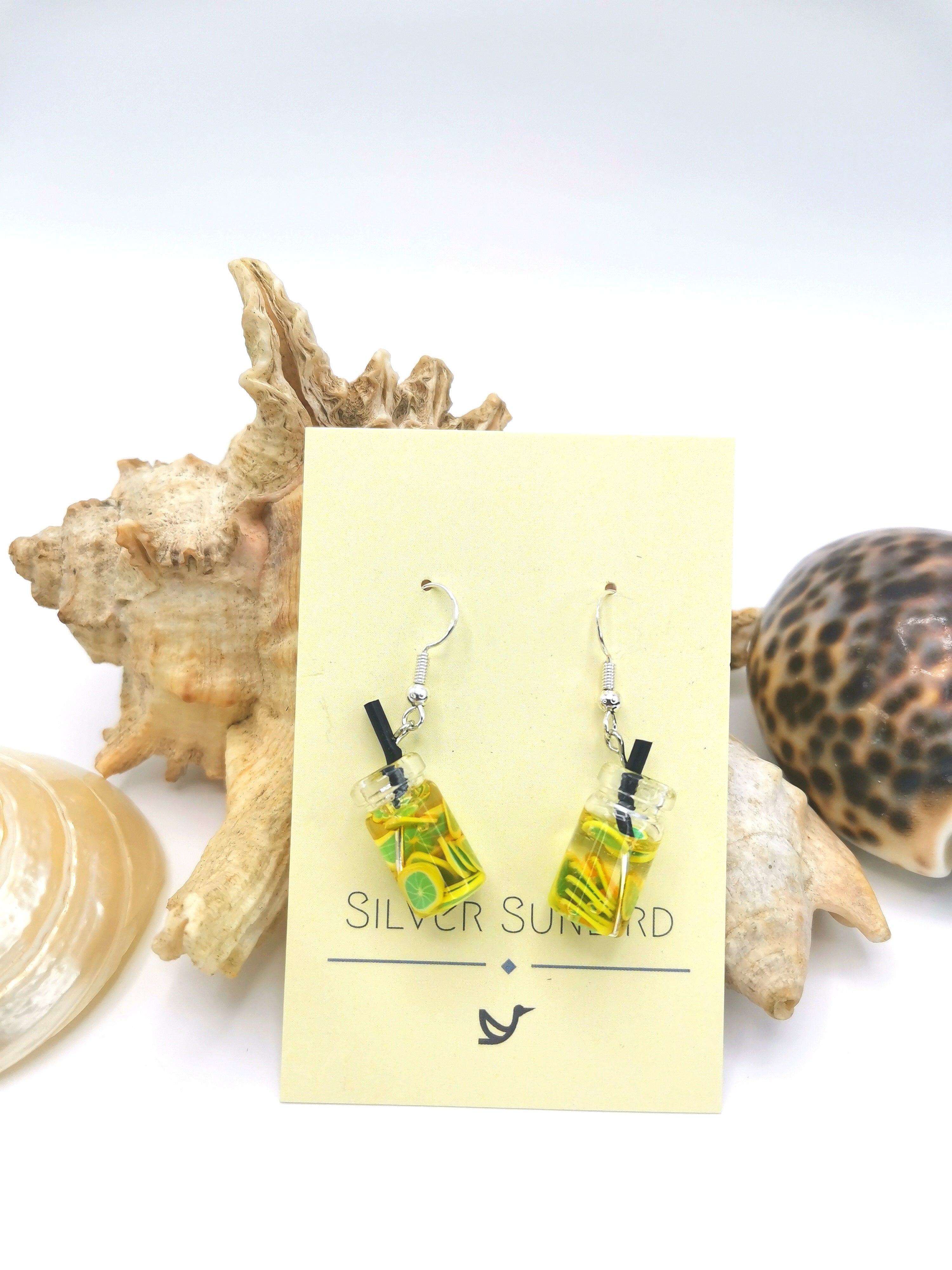 The Party Cocktail Earrings by Silver Sunbird are handmade with tiny glass bottles filled with yellow liquid. Displayed on a card against seashells and a white surface, they perfectly capture the essence of summer.