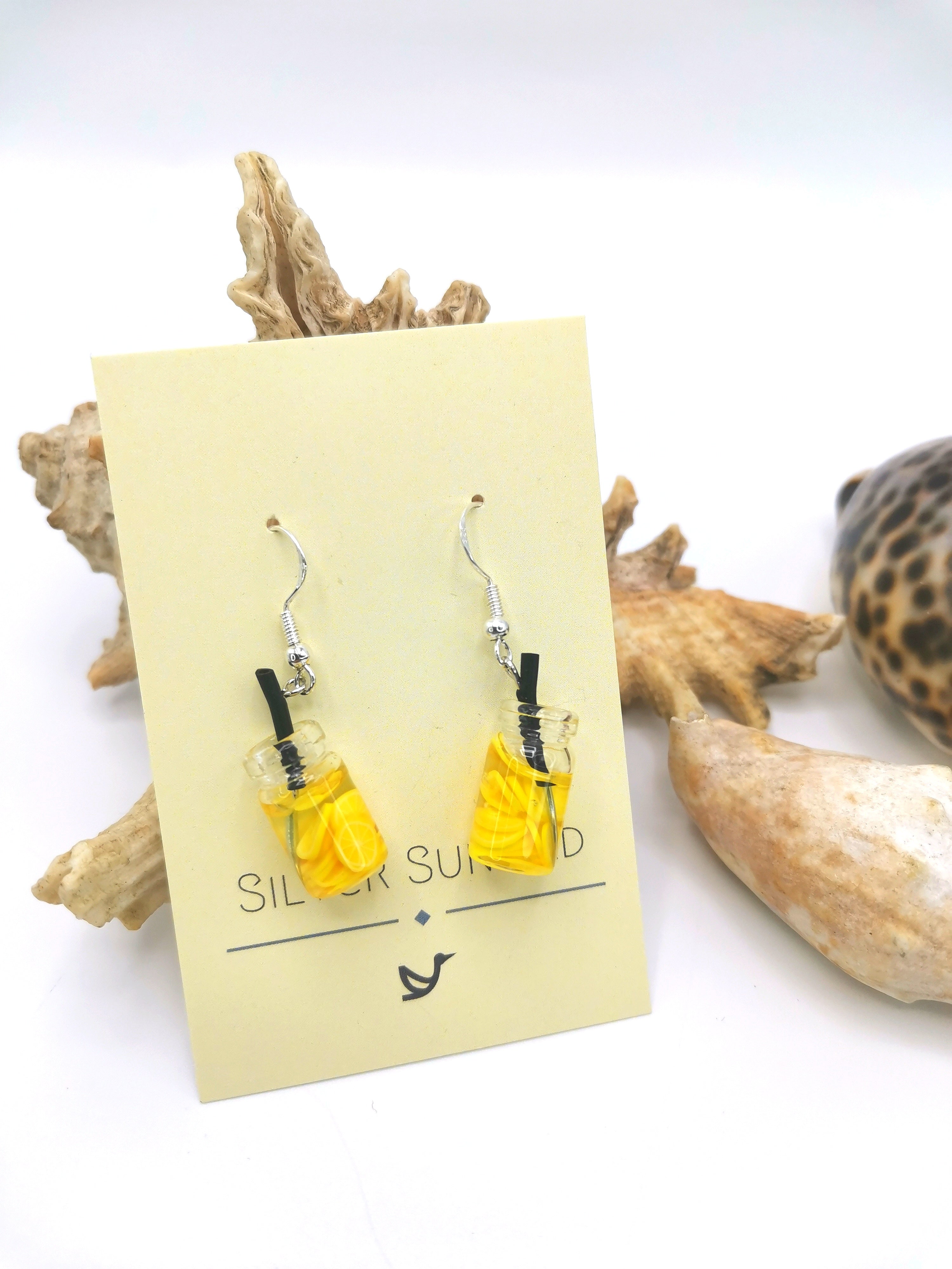 The Party Cocktail Earrings, by Silver Sunbird, are displayed on a card. These handmade earrings are shaped like transparent glasses filled with yellow liquid and black straws and are styled with seashells and driftwood to evoke summer vibes.