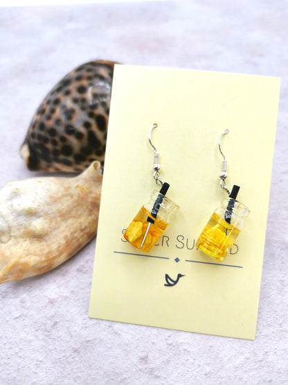 Two handmade sterling silver earrings, designed as small drinks with yellow liquid and black straws, hang on a card labeled &quot;Silver Sunbird.&quot; The Party Cocktail Earrings rest on a light-colored surface with seashells, perfectly capturing the essence of summer.